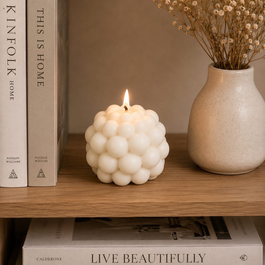 A white bubble-style candle is lit on a wooden shelf, styled with books and a ceramic vase with dried flowers against a warm beige wall.
شمعة بابل كريمي بتصميم فقاعات ثلاثية الأبعاد مودرن لتزيين طاولة القهوة والكونسول وديكور المنزل.