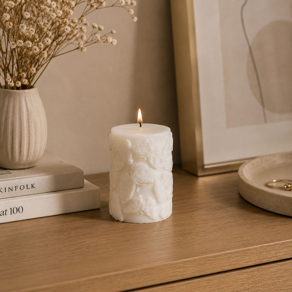 A lit white pillar candle with embossed floral relief details sits on a wooden table in a styled home setting, next to a vase of dried flowers and books.
شمعة ديكور كريمي أسطوانية مزينة بفراشات بارزة ثلاثية الأبعاد، مناسبة للكونسول وطاولة القهوة وديكور المنزل.