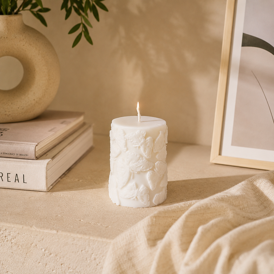 A lit white pillar candle with embossed classical figures, styled on a beige stone surface with books, a ceramic vase, and a framed print in a warm, minimalist interior.
شمعة ديكور كريمي أسطوانية مزينة بفراشات بارزة ثلاثية الأبعاد، مناسبة للكونسول وطاولة القهوة وديكور المنزل.