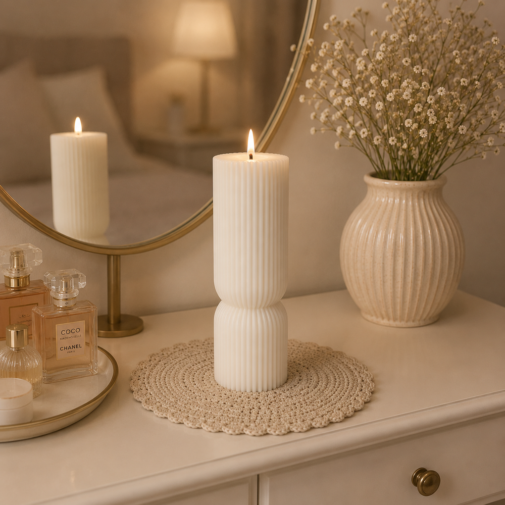 A tall white ribbed pillar candle stands lit on a round woven placemat on a cream dresser, with a matching cream ribbed vase of baby's breath and perfume bottles nearby. A round mirror reflects the candle in a softly lit bedroom setting.
شمعة عمود كريمي طويلة بتصميم مخطط هندسي مودرن، بارتفاع 16 سم، موضوعة على تسريحة بجوار مرآة وفازة ديكور.