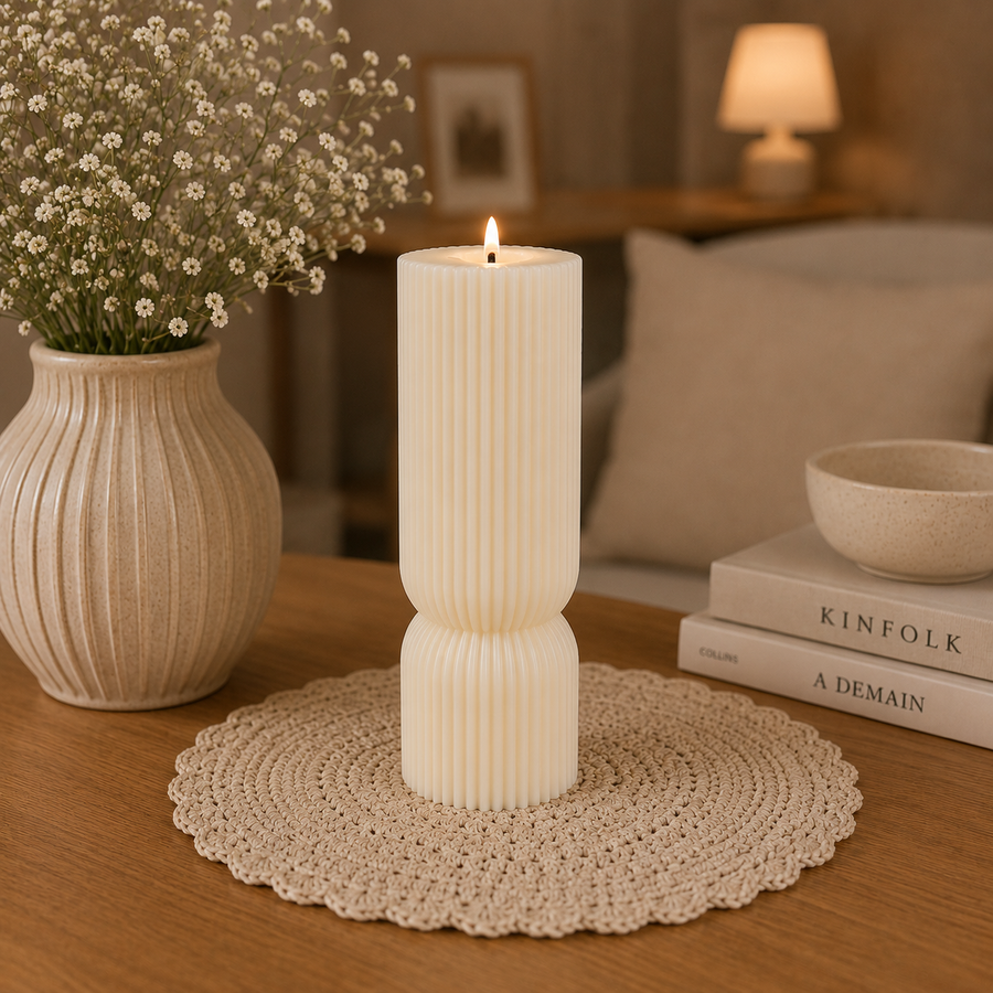 A ribbed cream-colored candle holder vase with a lit candle inside, styled on a wooden table with a beige crochet doily, a vase of baby's breath, and books in the background.
شمعة عمود كريمي طويلة بتصميم مخطط هندسي مودرن، بارتفاع 16 سم، موضوعة على تسريحة بجوار مرآة وفازة ديكور.