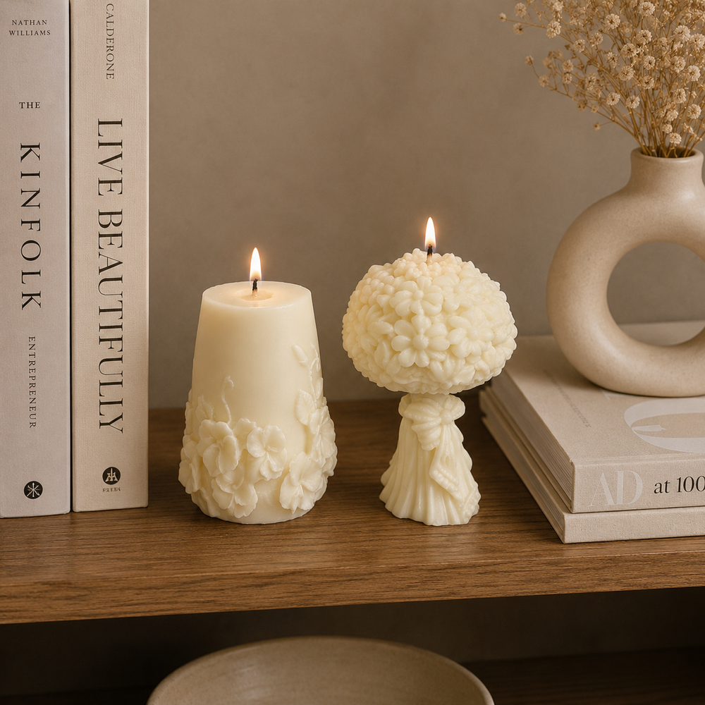 Two decorative white candles lit on a wooden shelf, styled with books, a ceramic vase with dried flowers, and a bowl against a warm beige wall.
طقم شموع ديكور ورد كريمي من قطعتين بتصميم زهور بارزة وباقة ورد لتزيين الكونسول والأرفف.