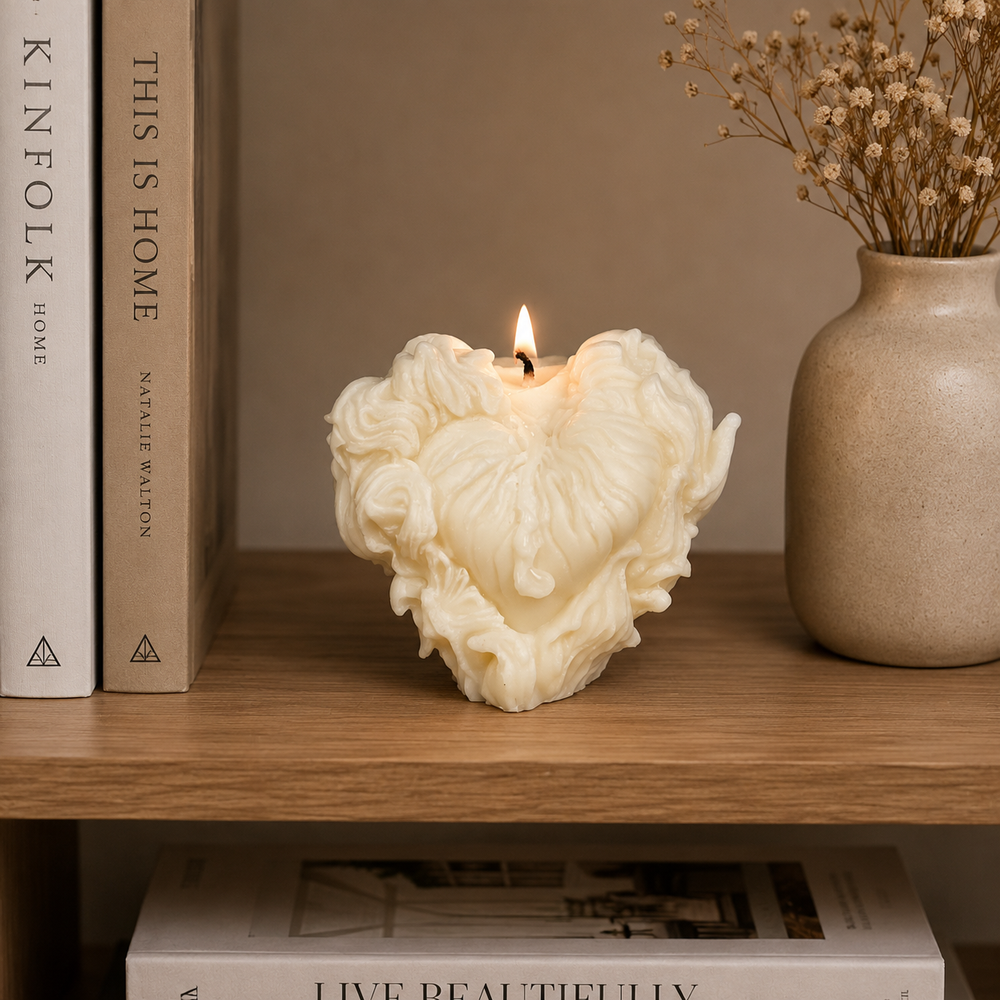 A sculpted cream-colored heart-shaped candle with a wick and a small flame, styled on a wooden shelf next to books and a ceramic vase with dried flowers.
شمعة قلب كريمي بتصميم فني ثلاثي الأبعاد وتفاصيل بارزة لتزيين الكونسول والأرفف وديكور المنزل المودرن.