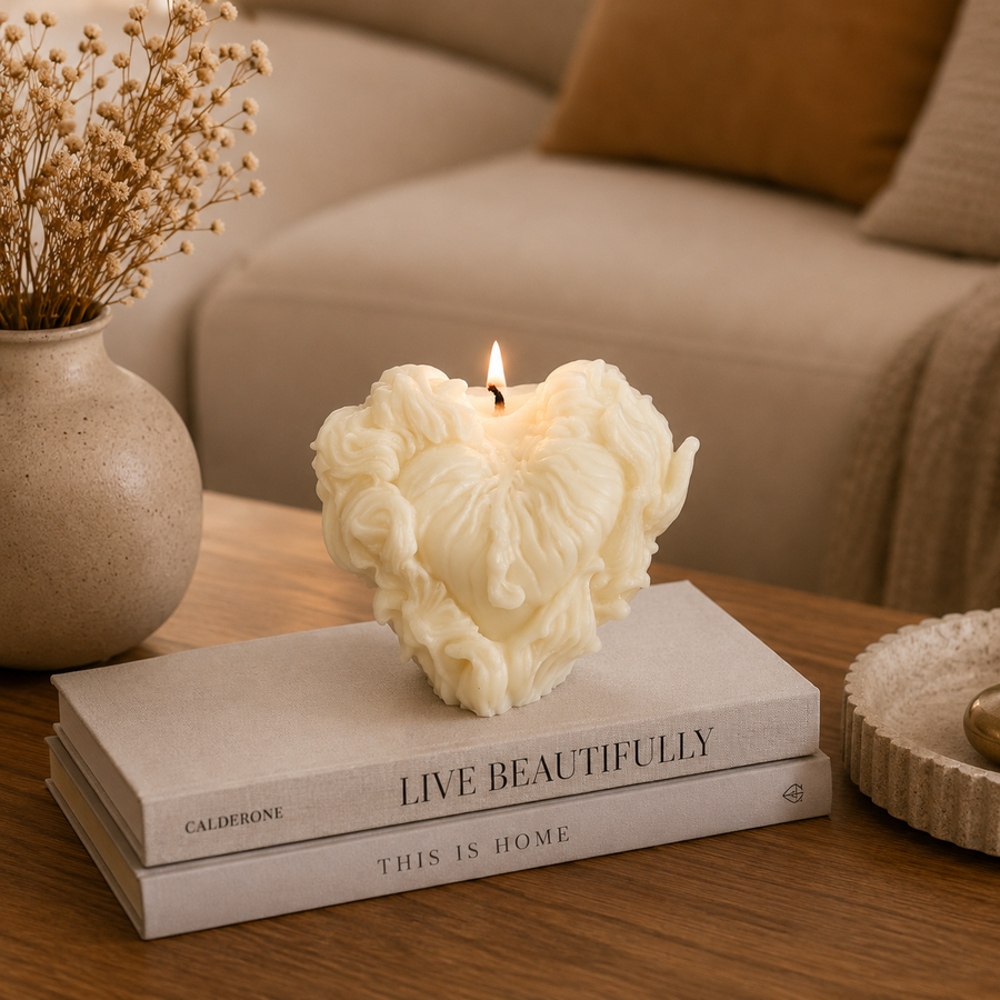 A sculptural cream-colored heart-shaped candle with a wavy, cloud-like surface sits lit on top of two stacked books on a wooden table. The setting is a cozy living room with a beige sofa, a vase of dried flowers, and a throw pillow in the background.
شمعة قلب كريمي بتصميم فني ثلاثي الأبعاد وتفاصيل بارزة لتزيين الكونسول والأرفف وديكور المنزل المودرن.