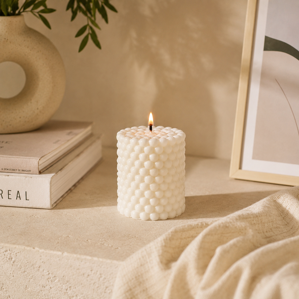 A lit white textured candle with a honeycomb-like bubble pattern sits on a beige tabletop in a warm, styled interior setting with books, a vase, and a framed print.
شمعة بابل كريمي بتصميم أسطواني وتفاصيل دائرية بارزة، مناسبة للكونسول وطاولة القهوة وديكور المنزل المودرن.