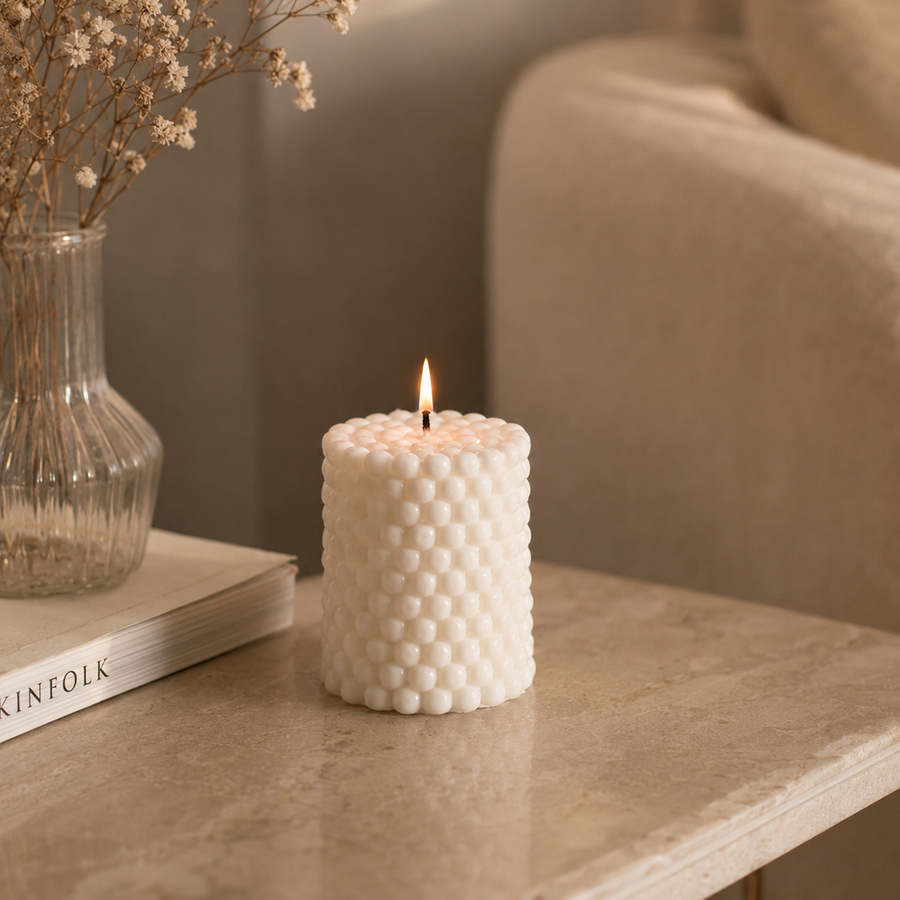 A lit white textured candle with a honeycomb-like bubble pattern sits on a beige stone table next to a glass vase with dried flowers and a book.
شمعة بابل كريمي بتصميم أسطواني وتفاصيل دائرية بارزة، مناسبة للكونسول وطاولة القهوة وديكور المنزل المودرن.