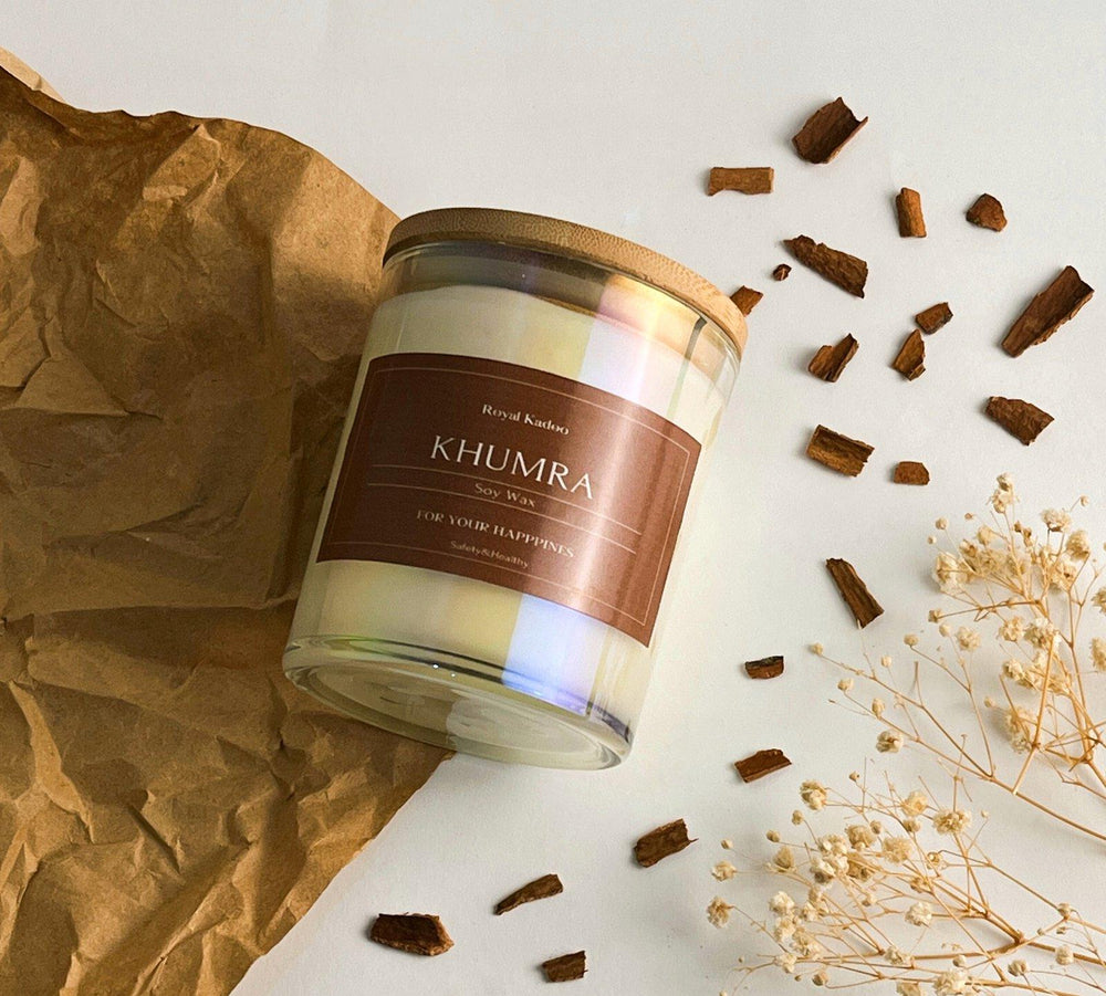Candle with 'Khumra' label on a white surface with brown paper and dried flowers.
شمعة خمرة معطرة من شمع الصويا داخل عبوة زجاجية بغطاء خشبي وملصق Khumra، منسقة بجوار قطع خشبية وزهور مجففة.