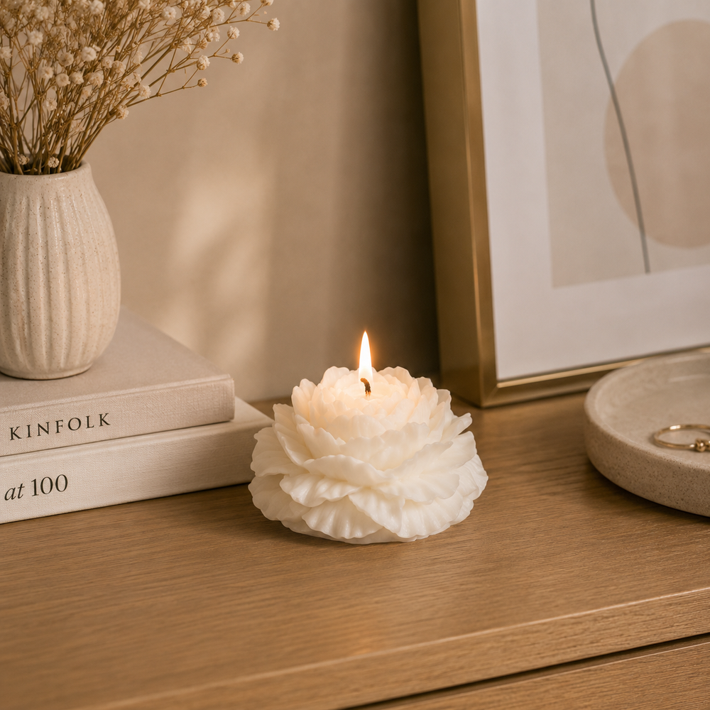 A white flower-shaped candle with a lit flame sits on a wooden tabletop in a styled interior setting, with books, a vase of dried flowers, and framed wall art nearby.
شمعة ديكور كريمي بتصميم زهرة الفاوانيا المتفتحة وبتلات بارزة، مناسبة لديكور المنزل والكونسول وطاولة القهوة والهدايا.