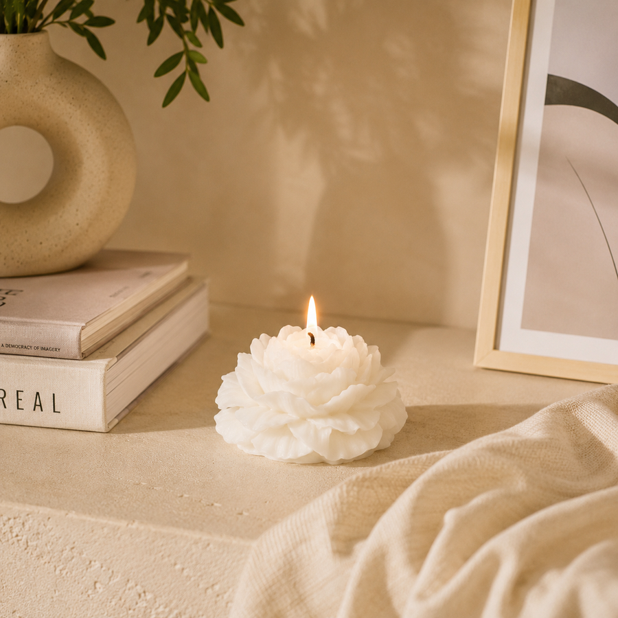 A white flower-shaped candle with a lit flame sits on a beige tabletop in a softly styled room scene with books, a vase, draped fabric, and framed wall art.
شمعة ديكور كريمي بتصميم زهرة الفاوانيا المتفتحة وبتلات بارزة، مناسبة لديكور المنزل والكونسول وطاولة القهوة والهدايا.