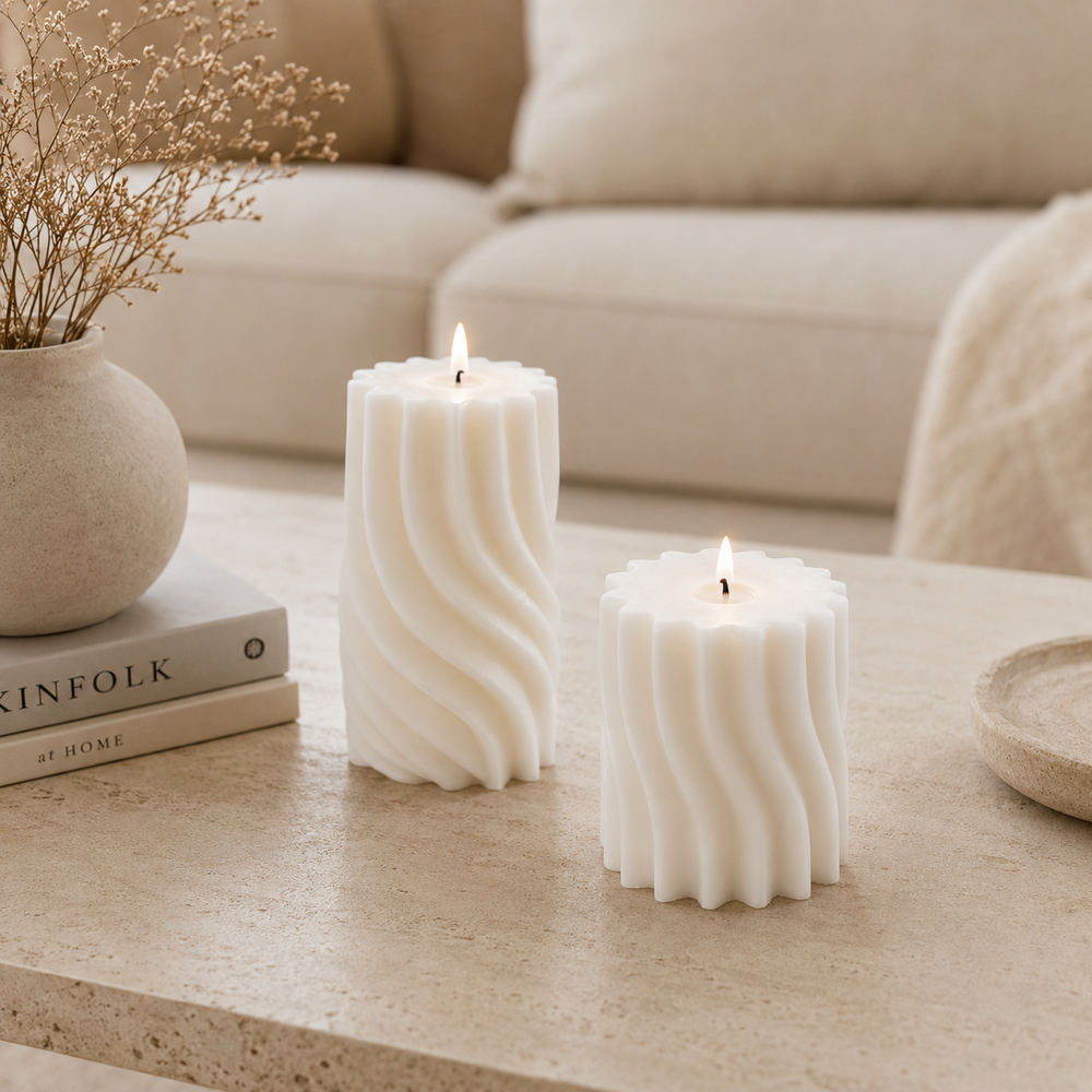 Two white ribbed pillar candles with lit flames sit on a beige stone table in a softly styled living room setting with a sofa, books, and a vase in the background.
طقم شمعتين كريمي بتصميم ويف انسيابي بأحجام مختلفة، مناسب لديكور طاولة القهوة والكونسول والأرفف المنزلية المودرن.