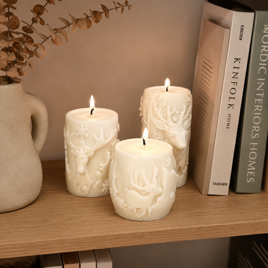 Three white pillar candles with embossed animal and floral designs, lit and displayed on a wooden shelf next to books and a ceramic vase.
طقم 3 شموع كريمي بتصميم الغزال الشتوي وتفاصيل بارزة بأحجام مختلفة لتزيين الكونسول وطاولة القهوة وديكور المنزل.