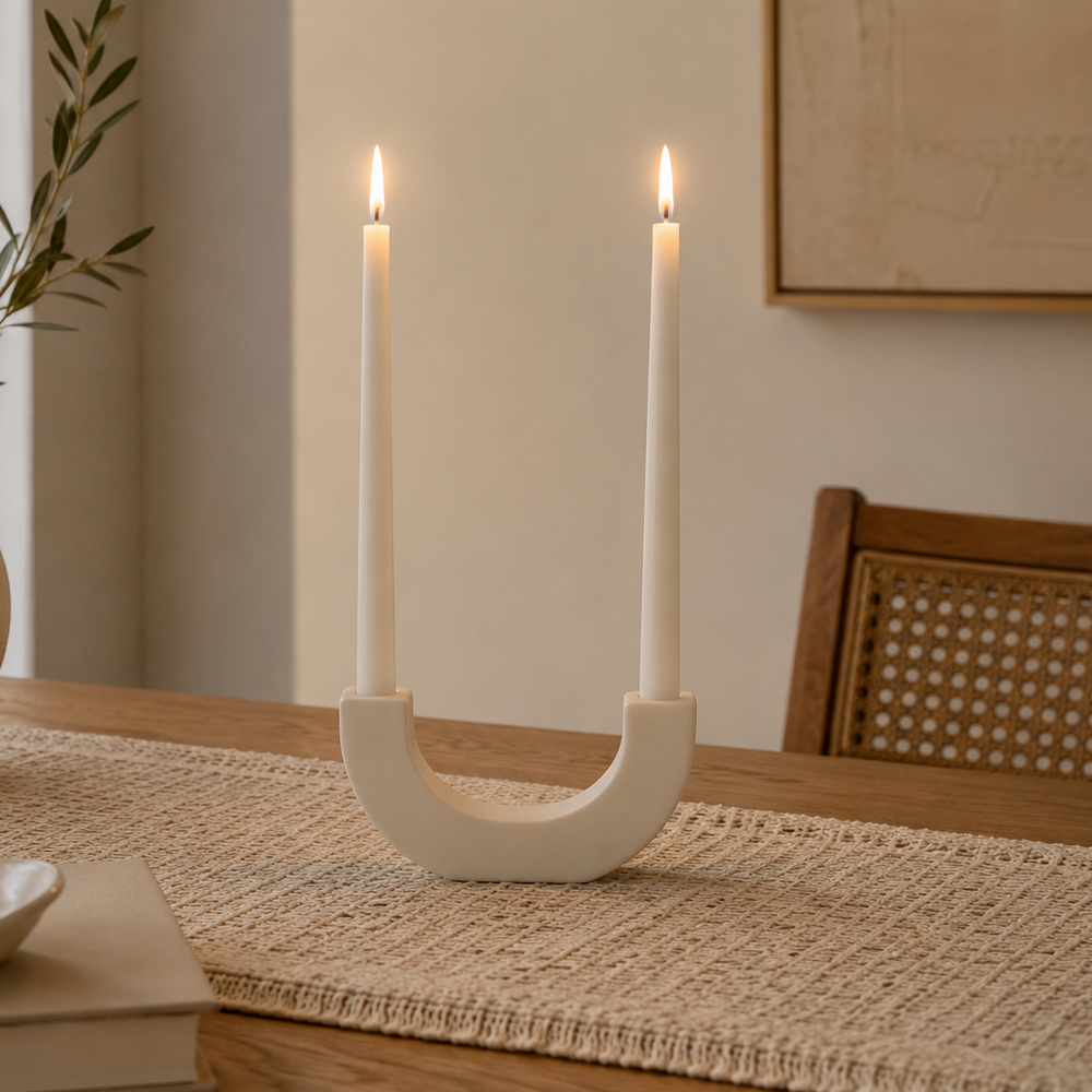 White U-shaped sculptural candlestick holder with two matching white taper candles, styled on a woven table runner on a wooden dining table in a modern dining room for elegant home décor and table centerpiece styling.