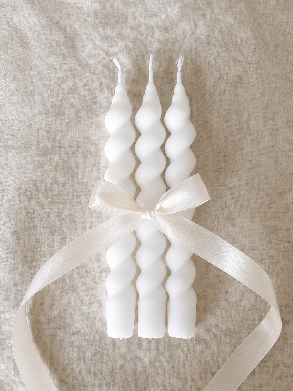 Set of white twisted taper candles tied with a cream satin ribbon, displayed on a textured beige fabric background for elegant home décor and gift styling.