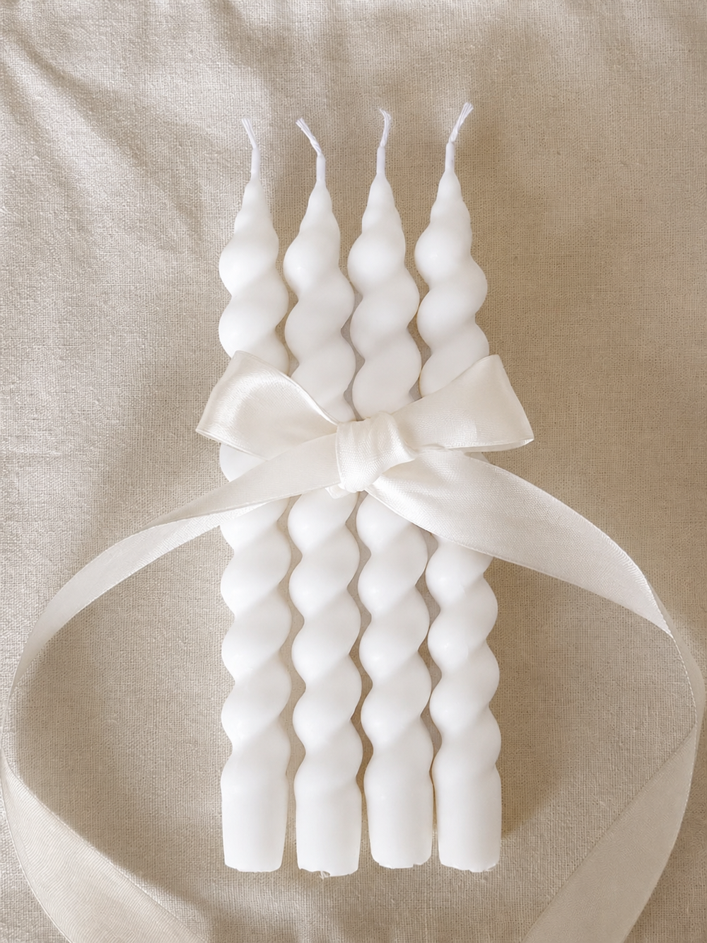 Set of four white twisted taper candles tied with a white satin ribbon, displayed on a textured beige fabric background for elegant home décor and gift presentation.